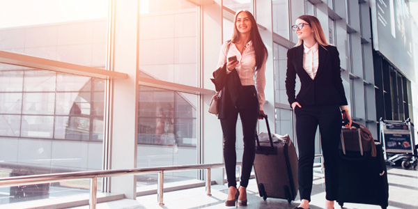 Airport setting. Two women dressed in business professional clothing walking with carry-ons.